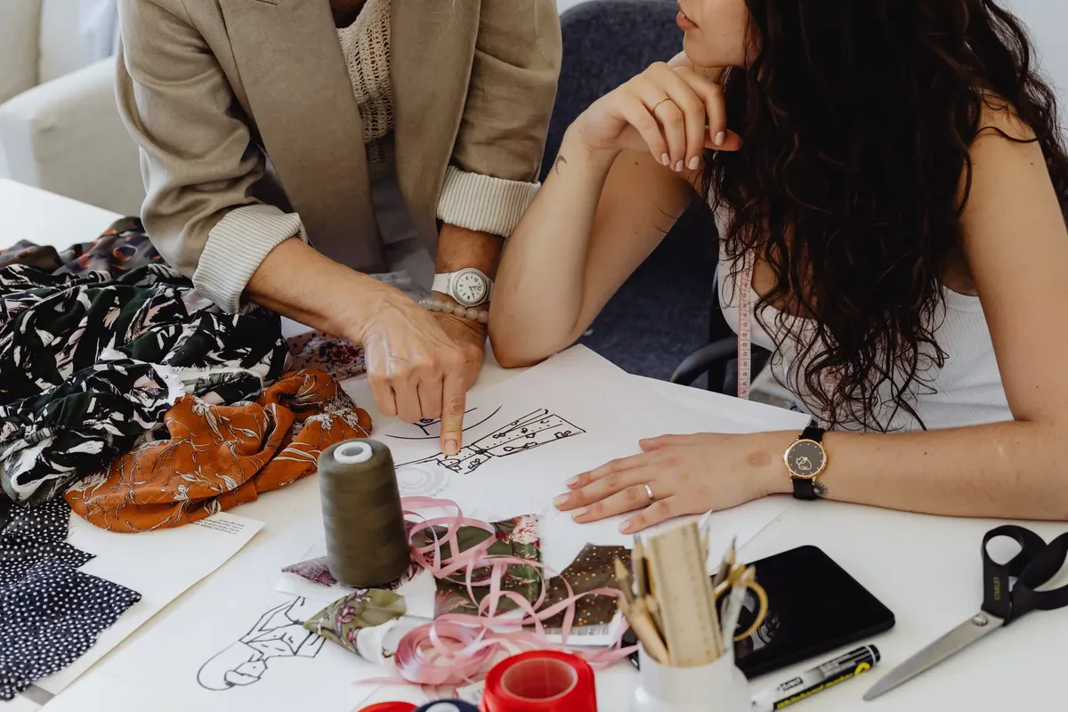 Two women collaborate on fashion sketches, fabrics, and tools, showcasing creative design process.
