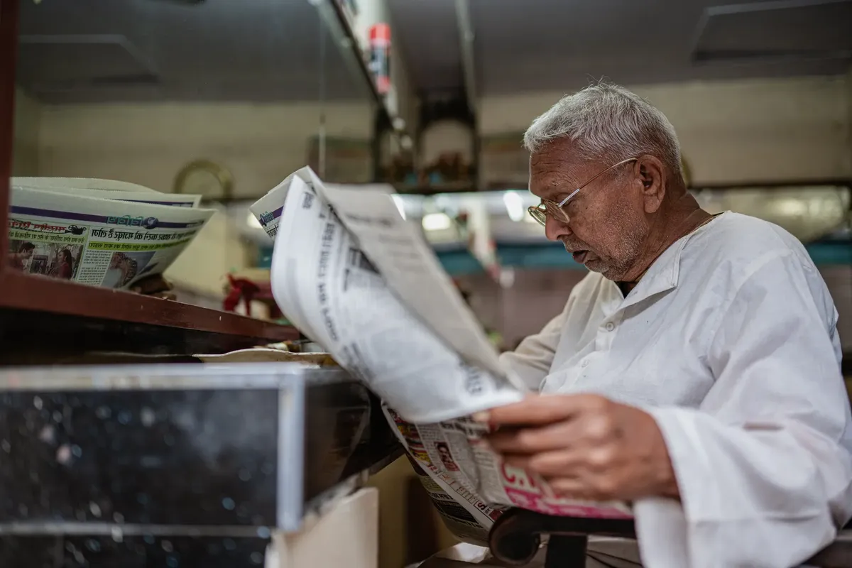 An elderly man in traditional attire reading a newspaper inside a shop in Varanasi, India.